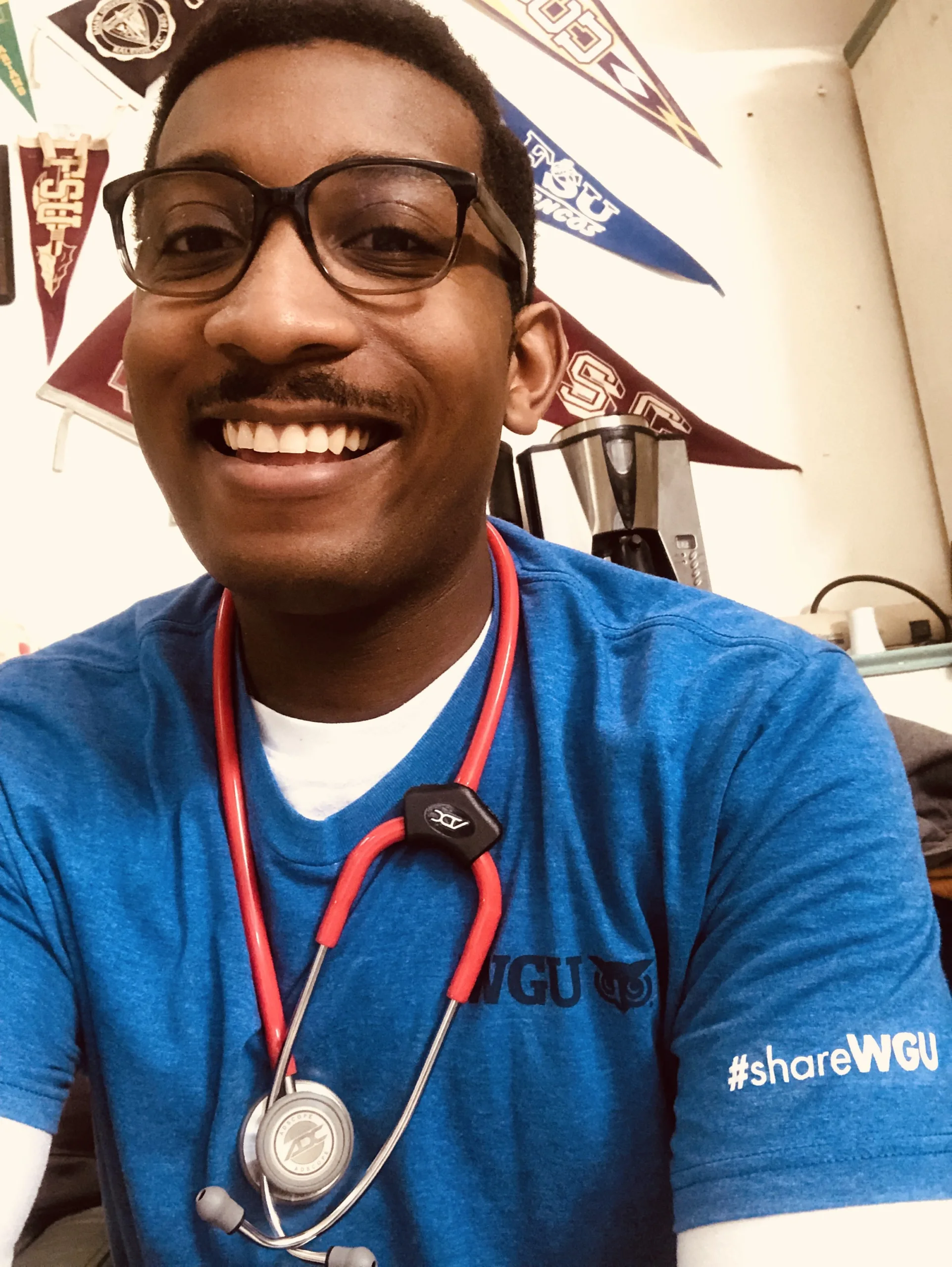 A person wearing glasses, a blue WGU shirt, and a red stethoscope smiles at the camera. School pennants hang on the wall in the background.