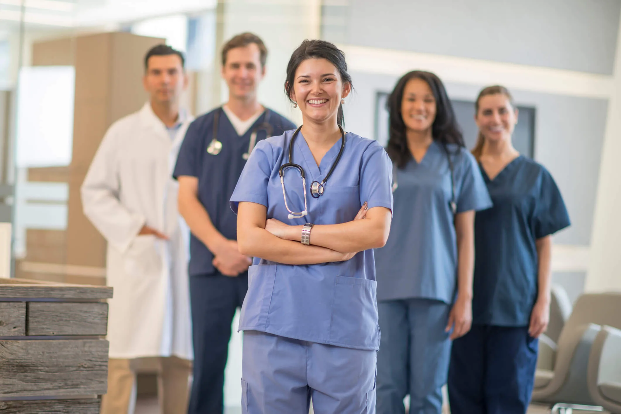 Five healthcare professionals in scrubs and lab coats stand together in a hospital setting, with a smiling woman in the foreground and four colleagues behind her.