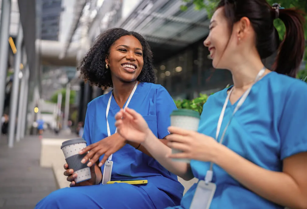 Two healthcare workers in blue scrubs sit outside, smiling and talking while holding takeaway coffee cups, with buildings and greenery in the background.