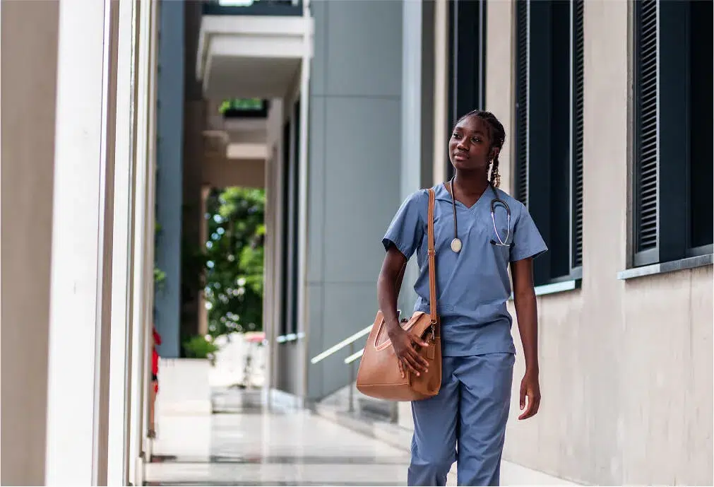 A nurse in blue scrubs with a stethoscope around her neck walks outdoors by a modern building, carrying a tan bag.
