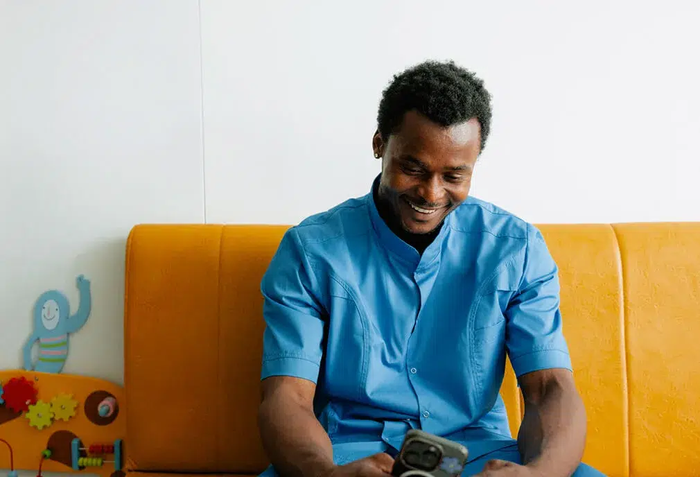 A man in blue scrubs sits on a yellow couch, smiling while looking at his phone. Colorful childrens toys are visible beside him.