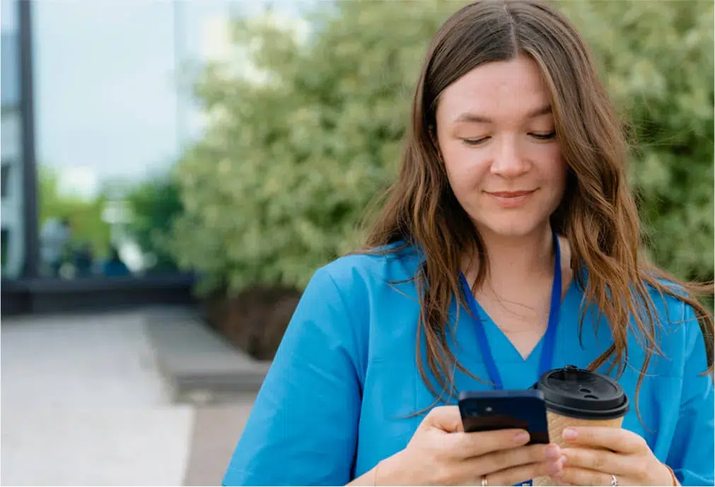 A woman in blue scrubs holds a coffee cup and looks at her smartphone outdoors, with greenery and a blurred building in the background.