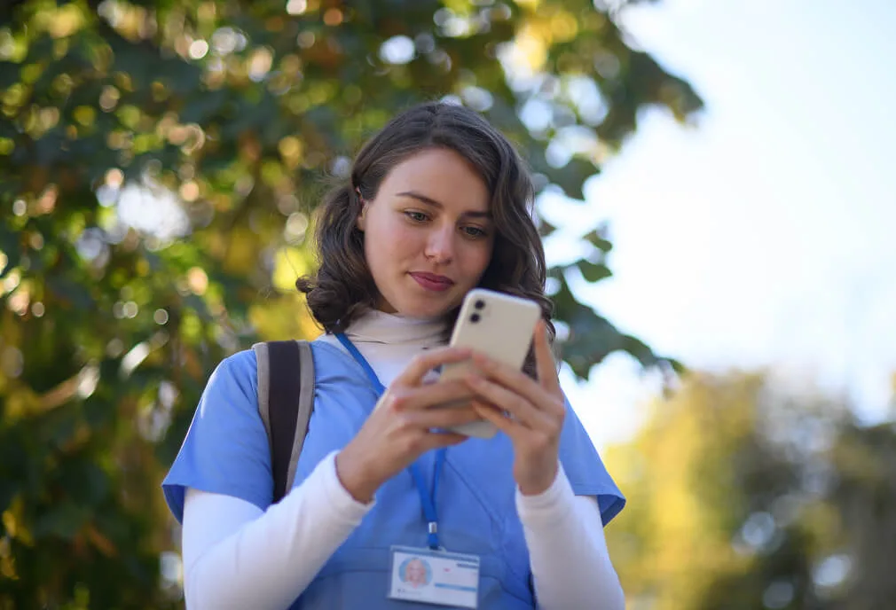 A woman in blue scrubs with a badge looks at her smartphone outdoors, with green trees and sunlight in the background.