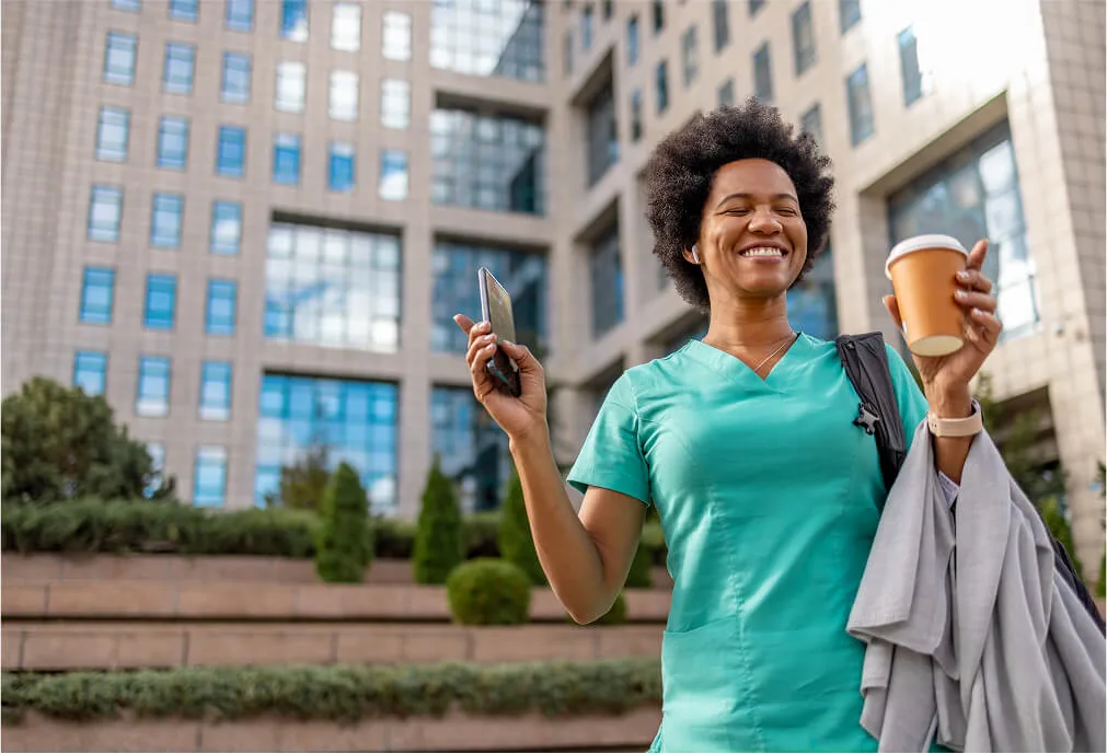 Smiling woman in green scrubs holds a phone and coffee cup outdoors, standing in front of a modern office building.