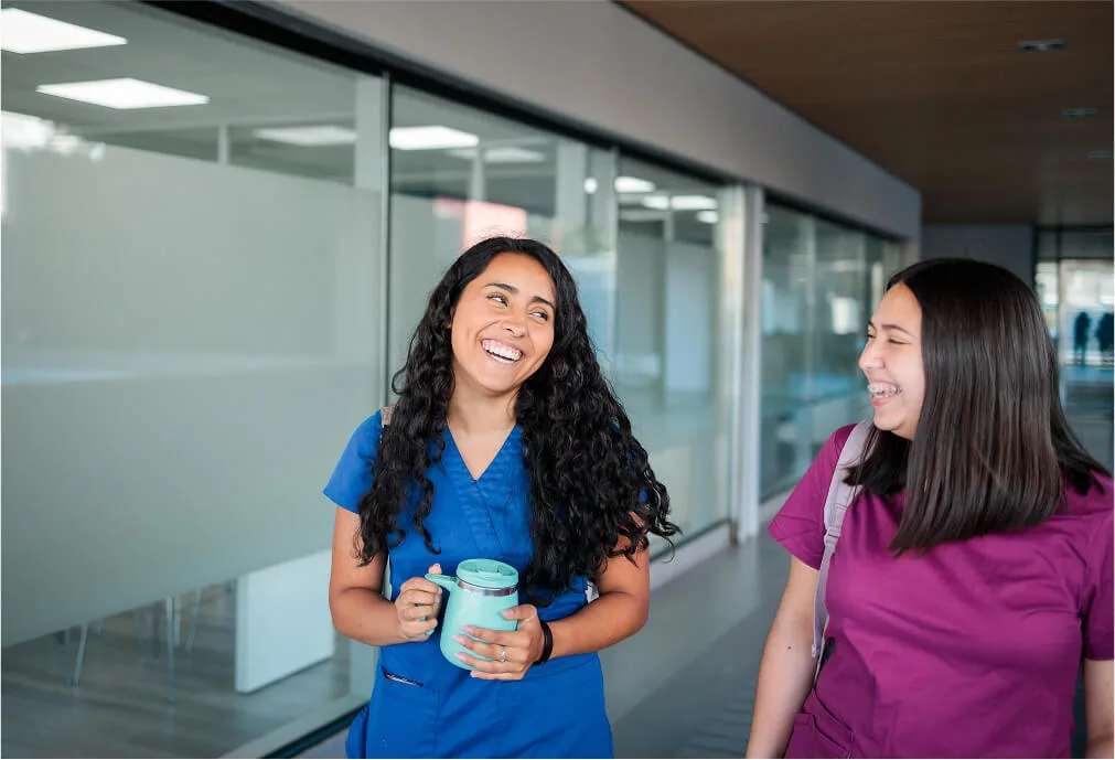 Two women in medical scrubs smiling and talking while walking indoors; one holds a mug.