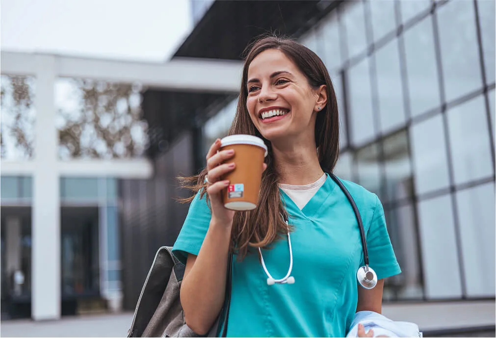 Smiling healthcare worker in teal scrubs, holding a coffee cup and bag, stands outside a modern building with glass walls.