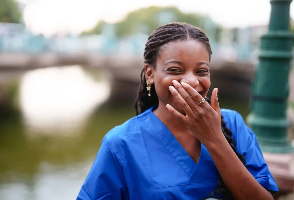 A woman in blue scrubs smiles and covers her mouth with her hand while standing outdoors near water.