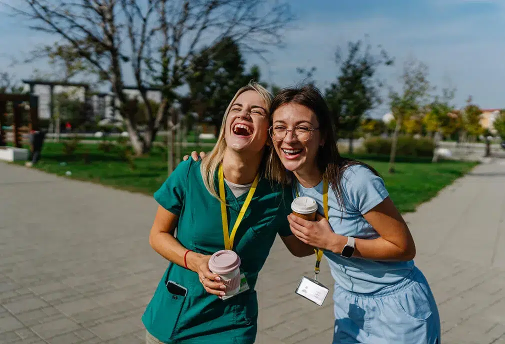 Two young women in scrubs with name badges laugh together outdoors, holding takeaway coffee cups and enjoying a sunny day in a park-like setting.