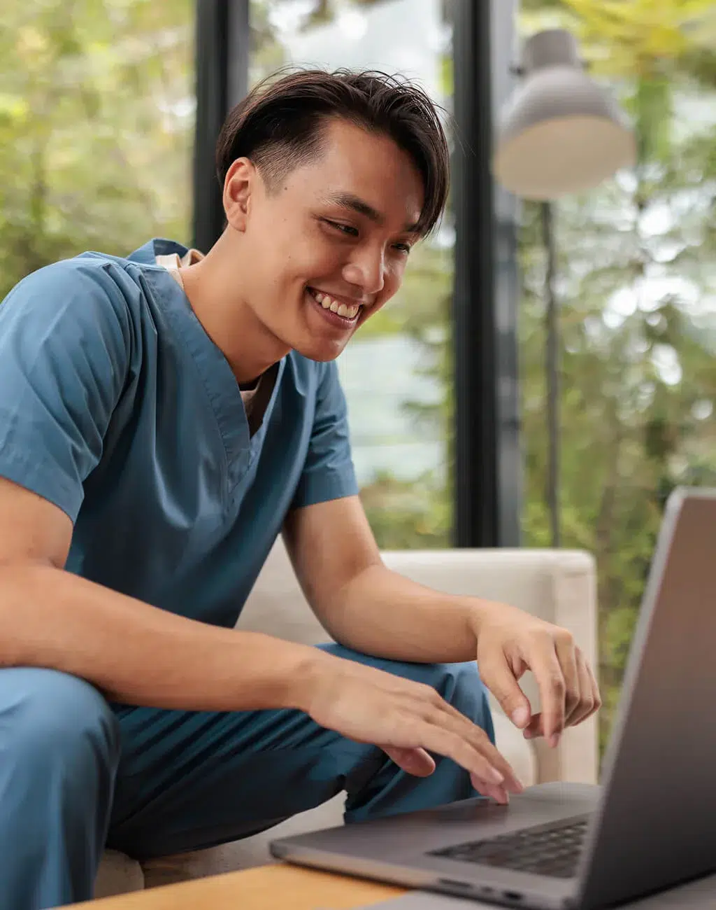 A smiling person in blue medical scrubs sits on a couch, typing on a laptop, with a large window and greenery in the background.