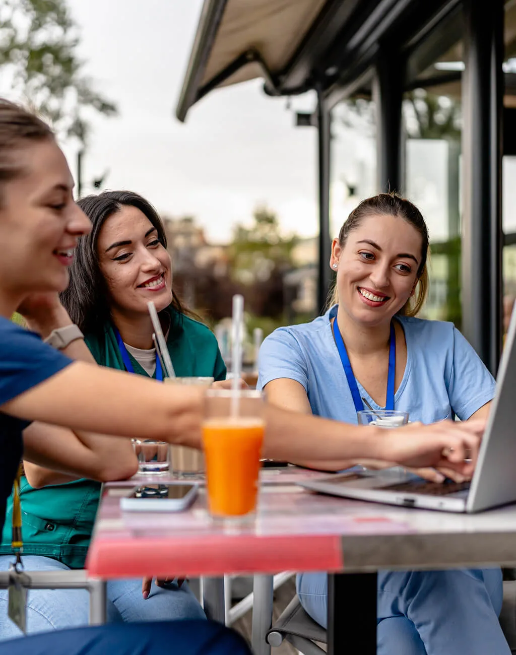 Three women in medical scrubs sit at an outdoor table, smiling and looking at a laptop, with drinks and notebooks in front of them.