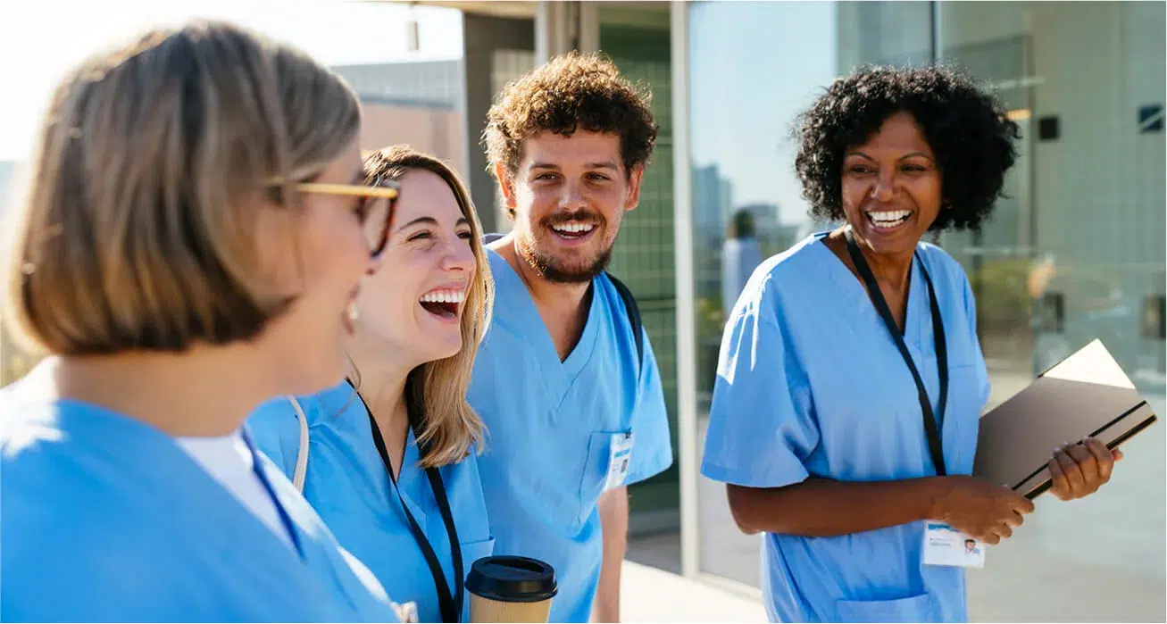 Four healthcare workers in blue scrubs are smiling and laughing together outside a modern building, appearing cheerful and engaged in conversation.