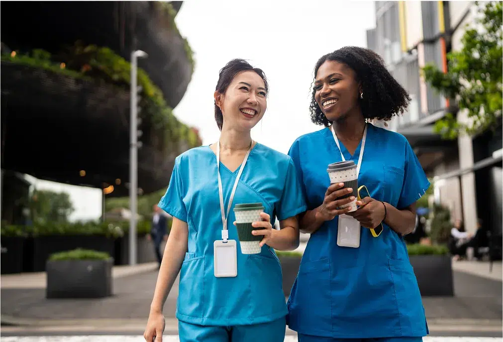 Two healthcare professionals in blue scrubs walk outside, smiling and holding coffee cups, with ID badges around their necks.