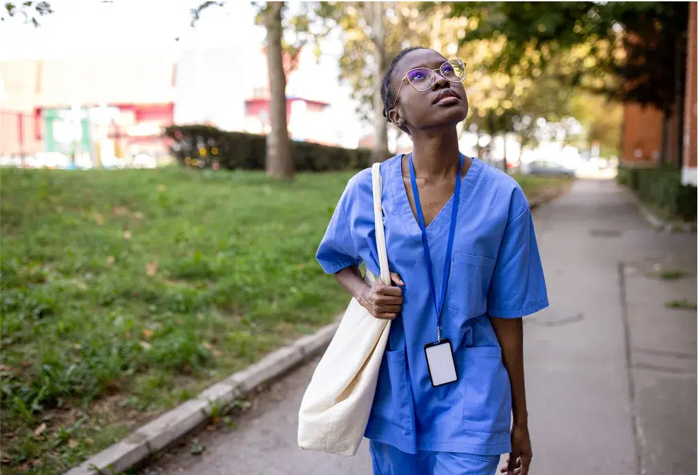 A person in blue medical scrubs and glasses walks outdoors on a sidewalk, carrying a tote bag and looking upward. Trees and grass line the path.