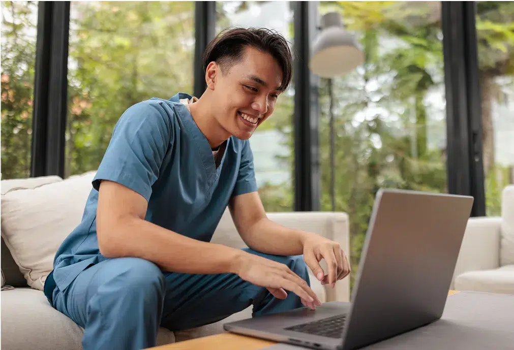 A smiling person in blue medical scrubs sits on a couch, using a laptop in a bright room with large windows and greenery outside.