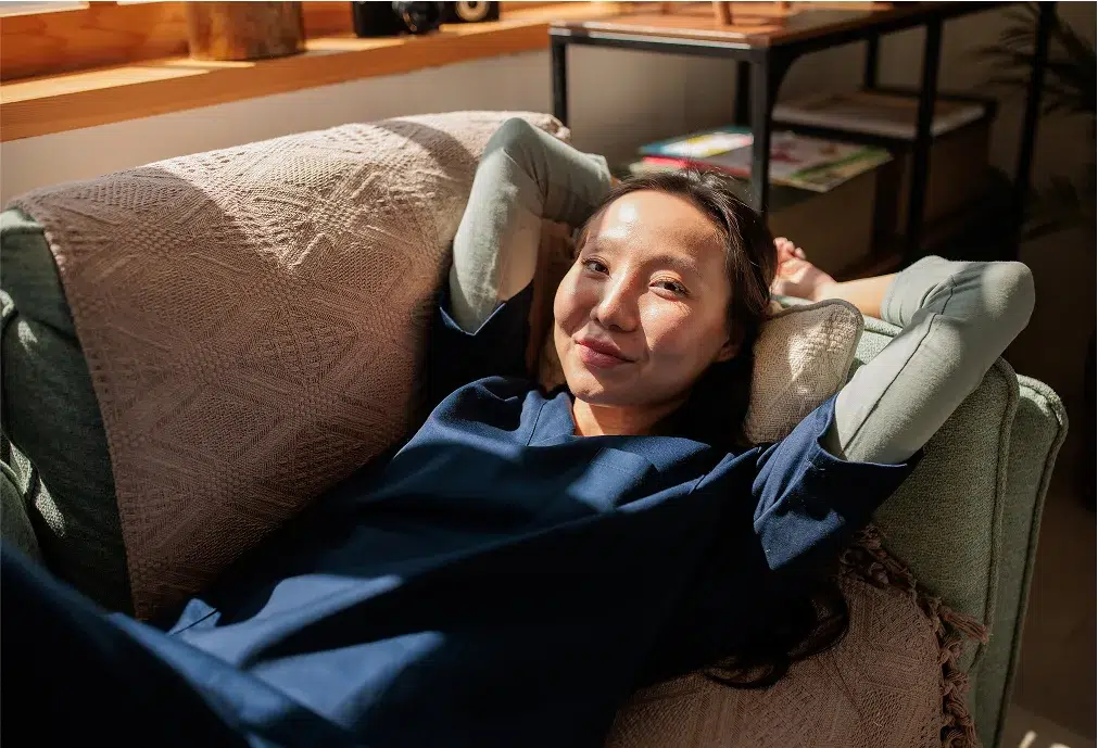 A woman in a blue outfit relaxes on a couch with her arms behind her head, smiling at the camera in a sunlit room.