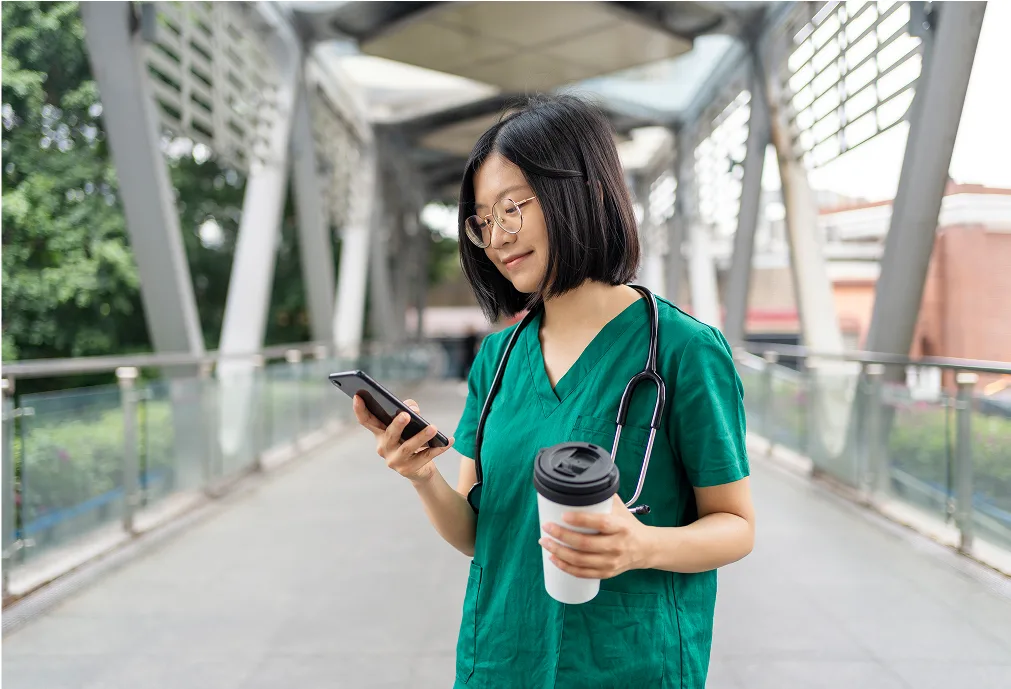 A woman in green scrubs with a stethoscope around her neck holds a coffee cup and looks at her phone while standing on a modern pedestrian bridge.