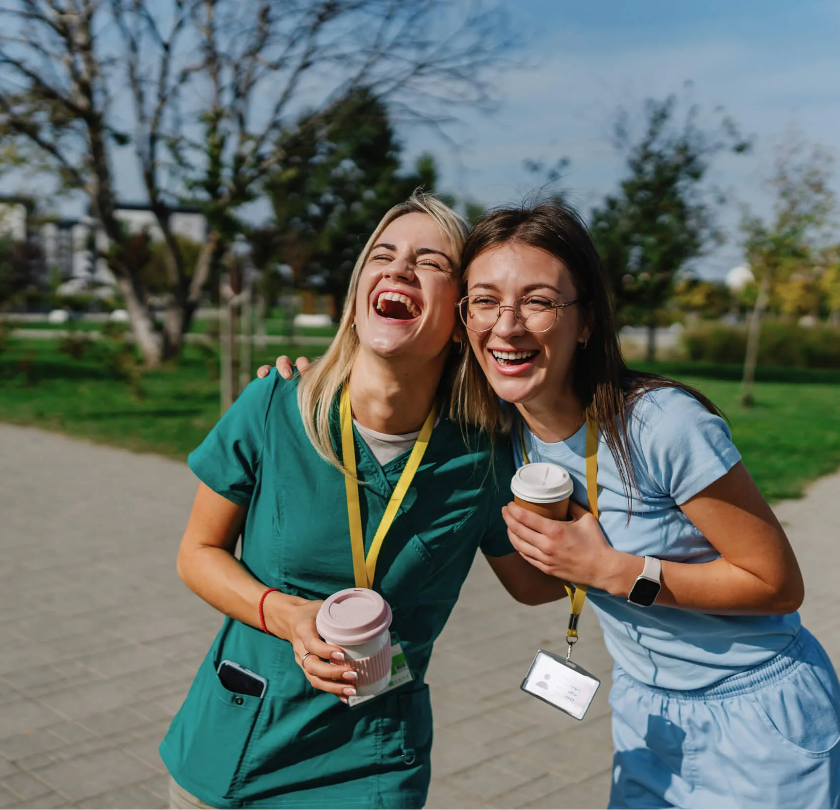 Two young women in scrubs, holding coffee cups and laughing together outdoors on a sunny day, wearing yellow lanyards and name badges.