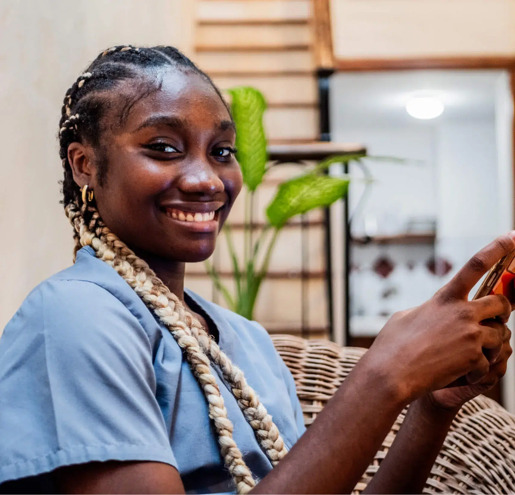 A woman with long braided hair sits on a wicker chair, smiling and holding a smartphone, with indoor plants and home decor in the background.