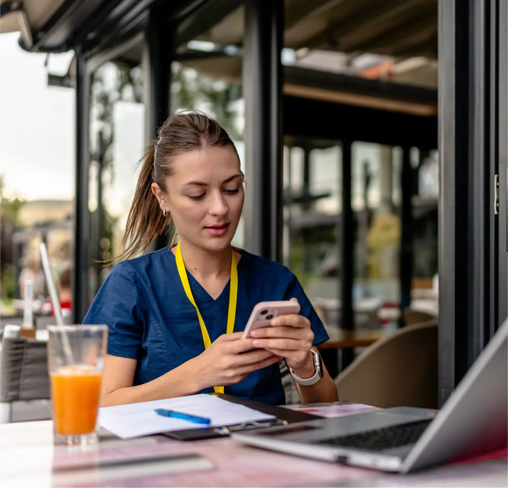 A person in scrubs with a yellow lanyard sits at an outdoor table, using a phone. Theres a laptop, paper, pen, and a glass of orange juice on the table.
