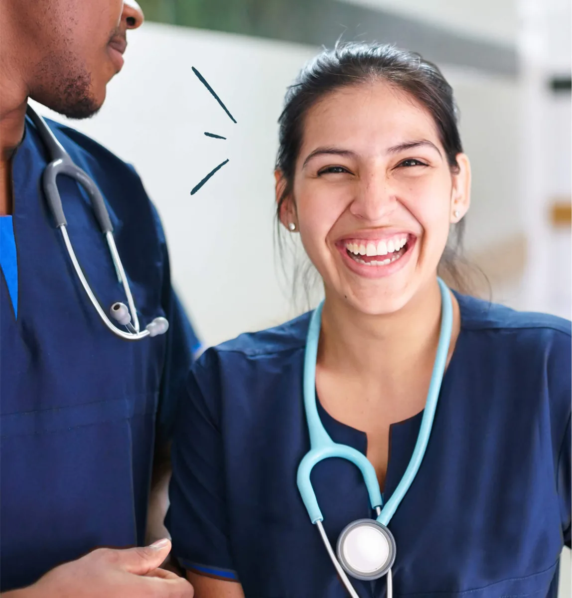 Two healthcare professionals in blue scrubs and stethoscopes, one smiling brightly at the camera, standing indoors.