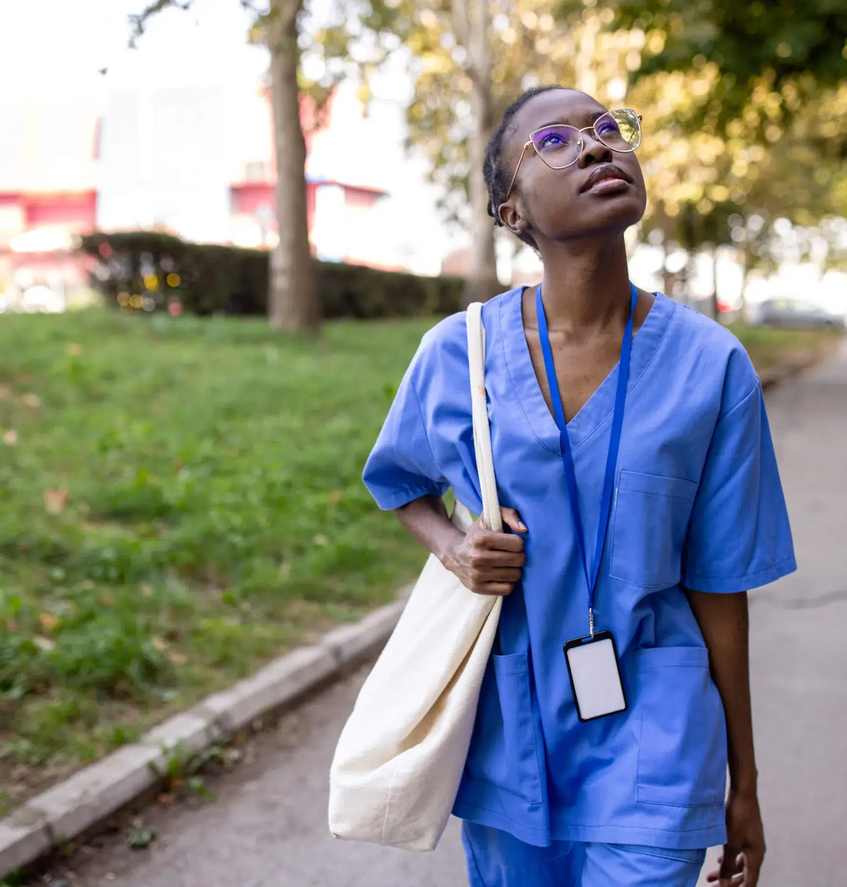 A healthcare worker in blue scrubs walks outdoors, carrying a cream tote bag and wearing a lanyard with an ID badge, looking up thoughtfully. Trees and grass are in the background.