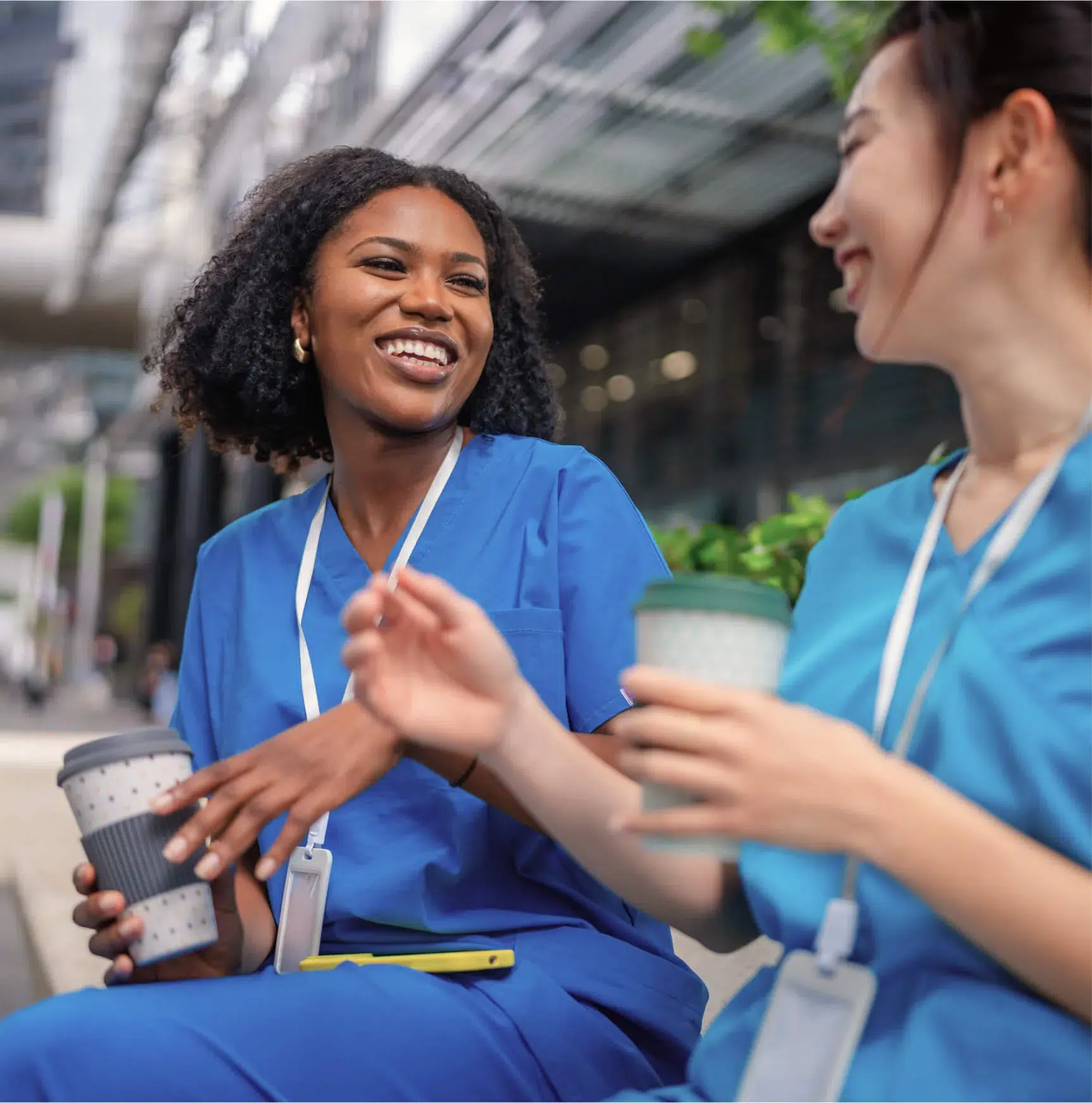Two women in blue scrubs sit outside, smiling and talking while holding coffee cups. Both wear ID lanyards, suggesting they are healthcare professionals on a break.