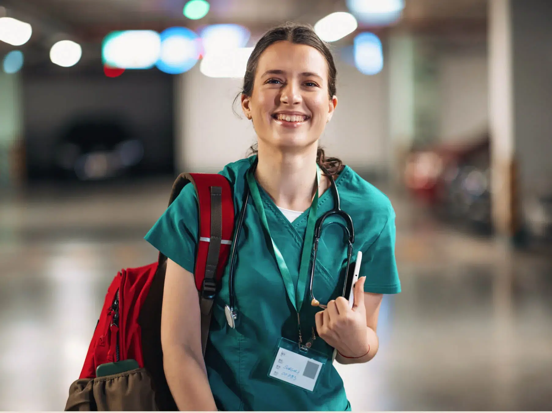 Smiling healthcare worker in green scrubs with a stethoscope and red backpack, standing in a brightly lit indoor setting.