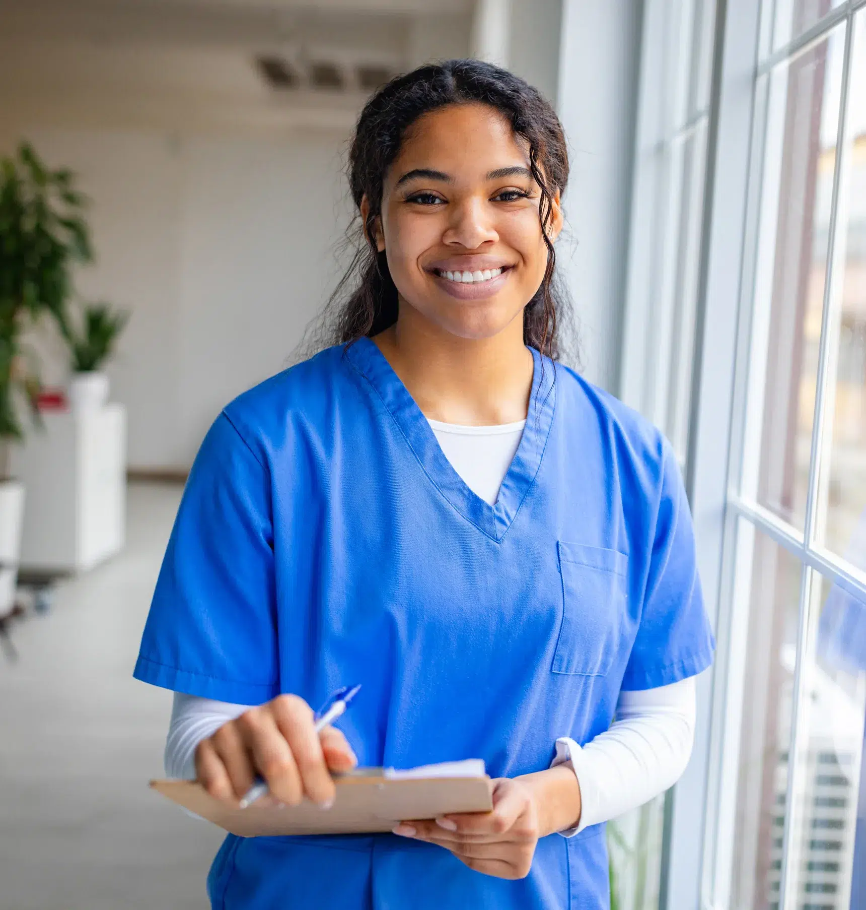 A nurse in blue scrubs stands by a window, smiling and holding a clipboard with a pen in hand.