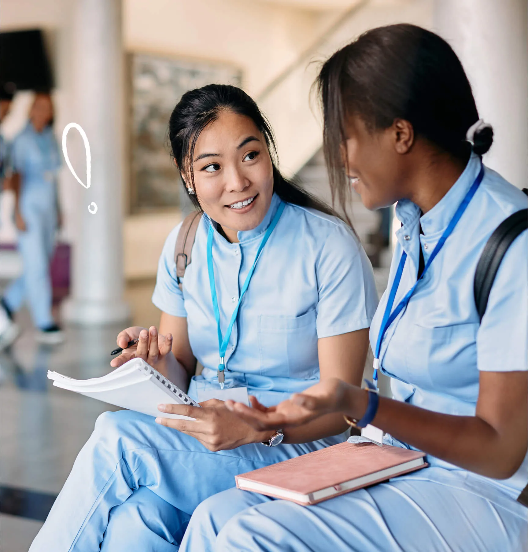 Two young women in light blue uniforms sit and talk, holding notebooks and wearing name tags, appearing engaged in conversation.