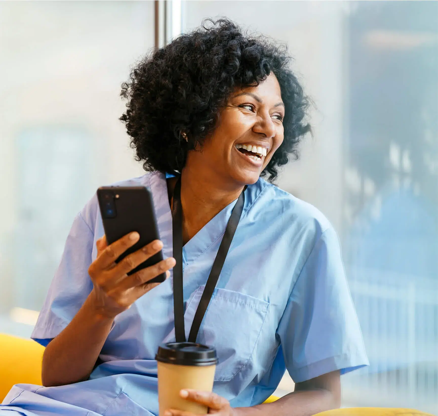 Smiling woman in medical scrubs holds a smartphone and a takeaway coffee cup, sitting indoors near a window.