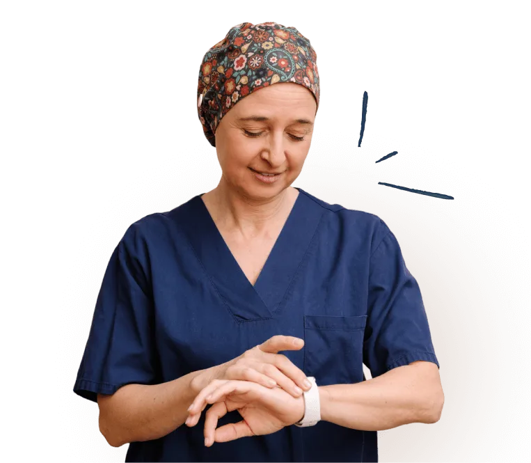 A healthcare worker in blue scrubs and a patterned cap looks down and touches a smartwatch on her wrist.