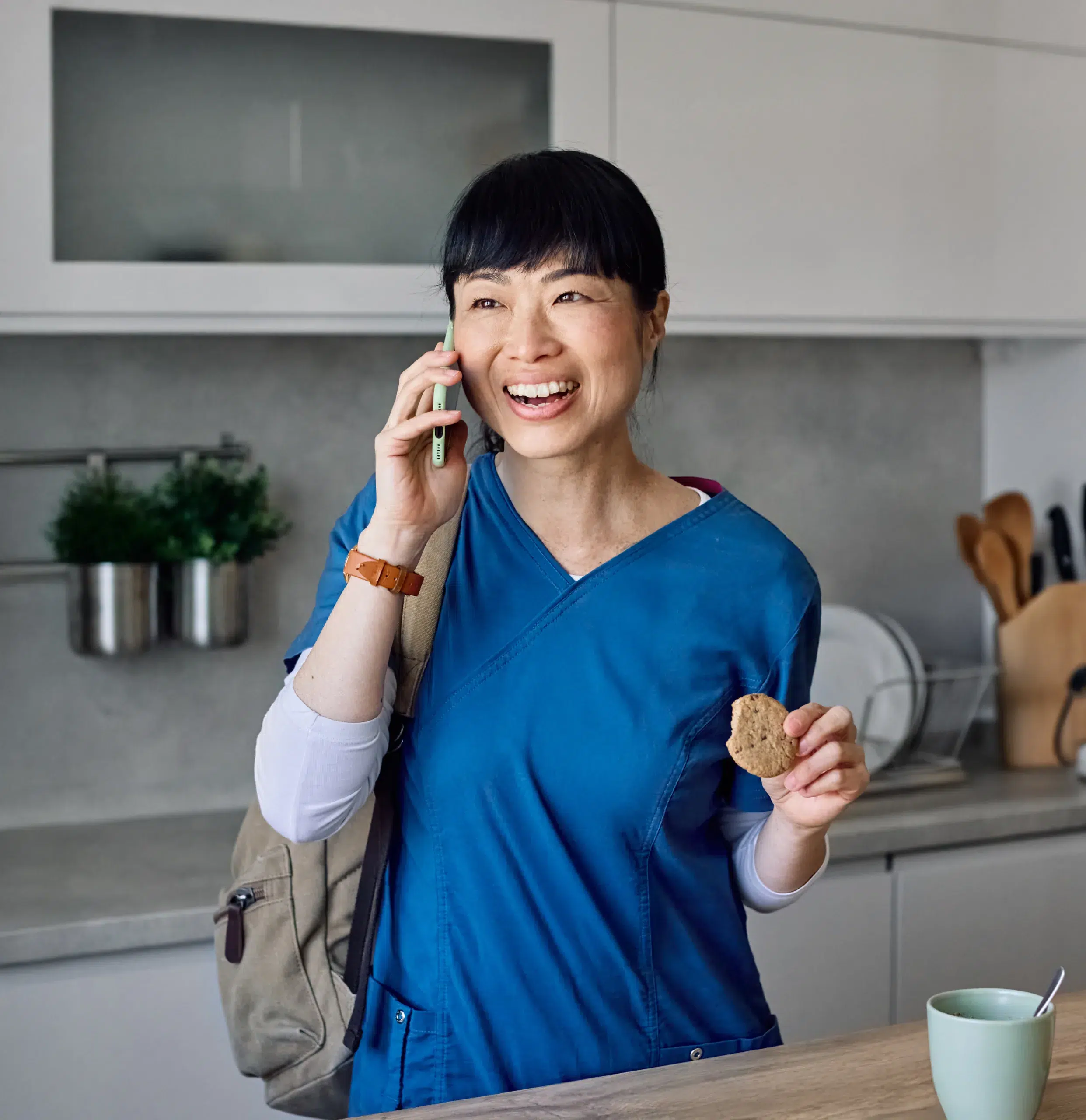 Smiling woman in blue scrubs talks on the phone in a kitchen, holding a cookie and a backpack—perhaps discussing the best calendar app for nurses.