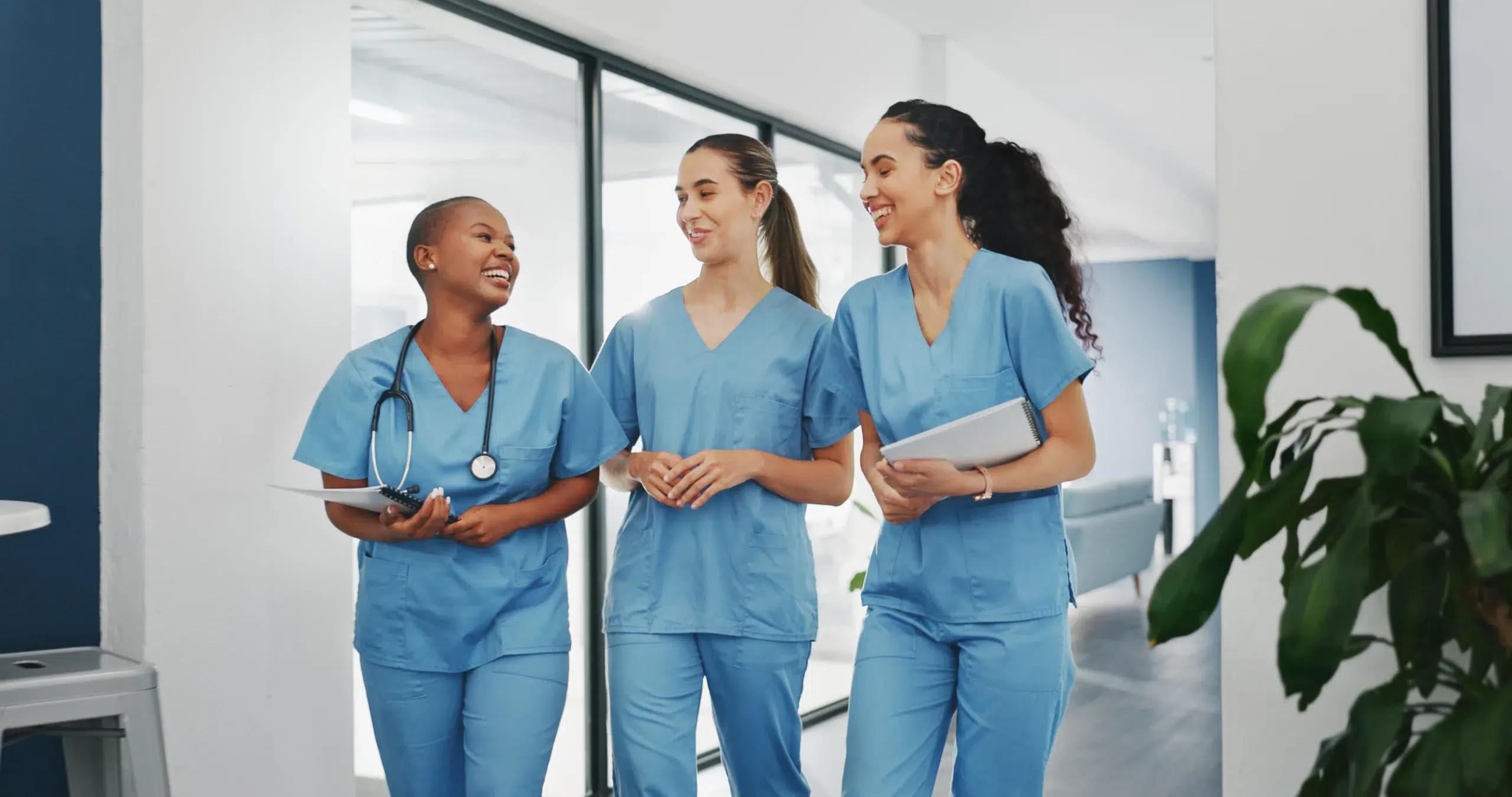 Three healthcare professionals in blue scrubs walk down a hallway, smiling and talking together about the best nursing schedule app, holding a tablet, clipboard, and stethoscope.