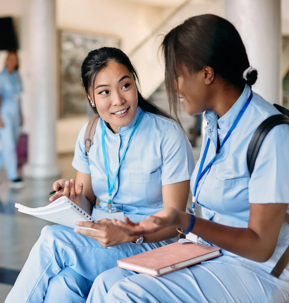 Two young women in light blue scrubs sit together indoors, discussing notebooks and nurse scheduling software, possibly students or healthcare professionals.