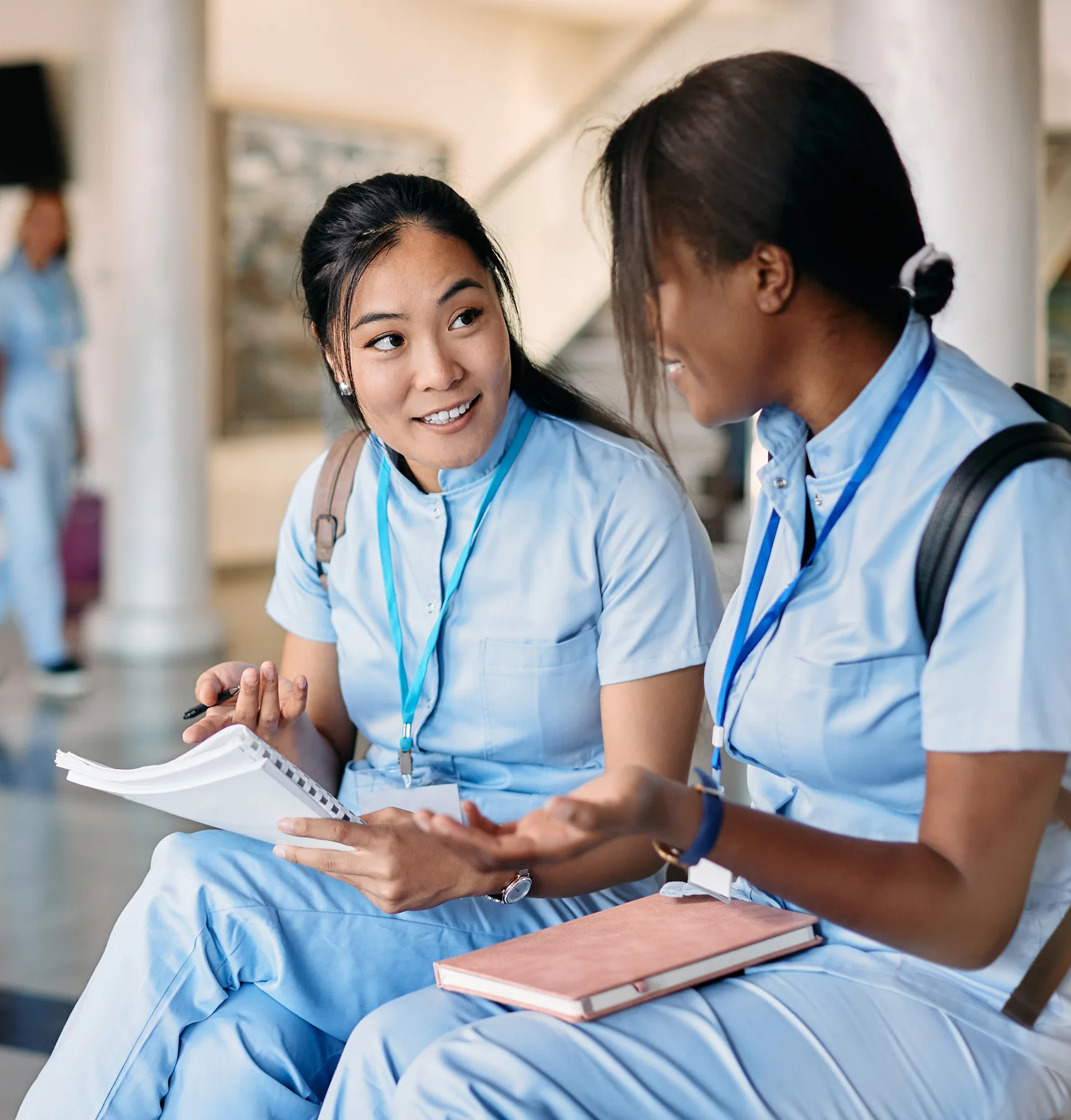 Two young women in light blue scrubs sit together indoors, discussing notebooks and nurse scheduling software, possibly students or healthcare professionals.