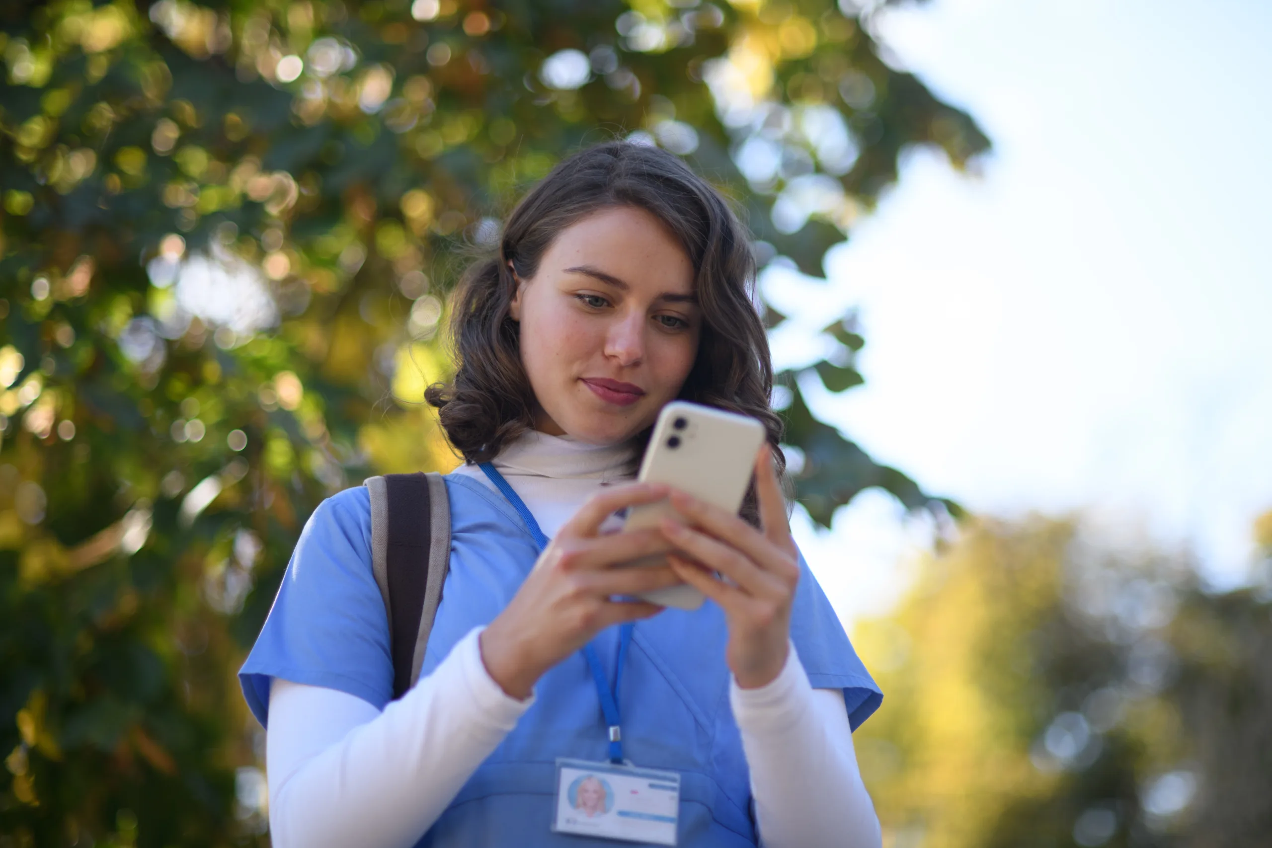 A woman in blue scrubs and a lanyard checks nurse scheduling software on her smartphone outdoors, with green trees blurred in the background.