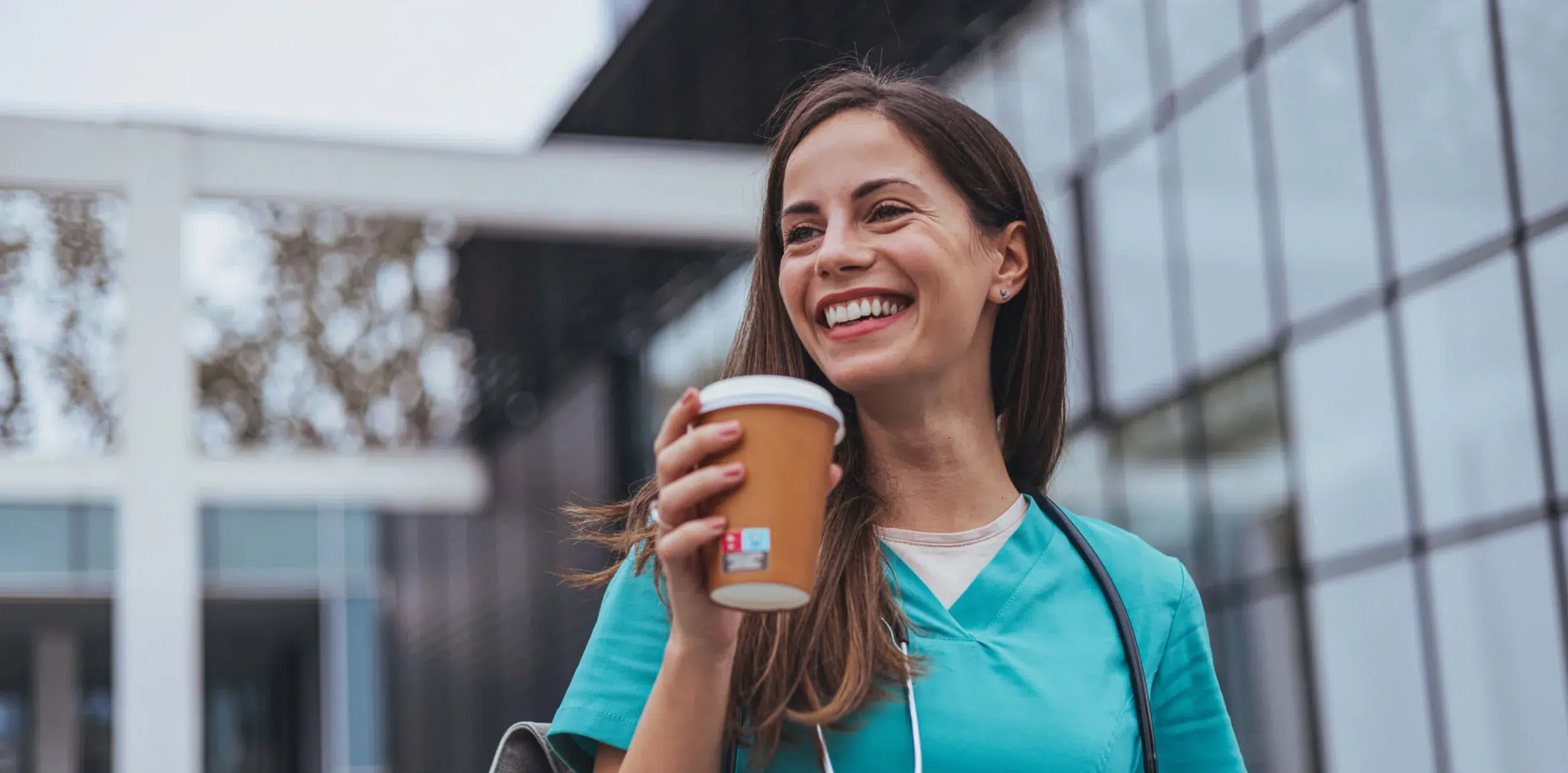 A smiling woman in teal scrubs holds a coffee cup outside a modern glass building, suggesting a healthcare professional on a break.