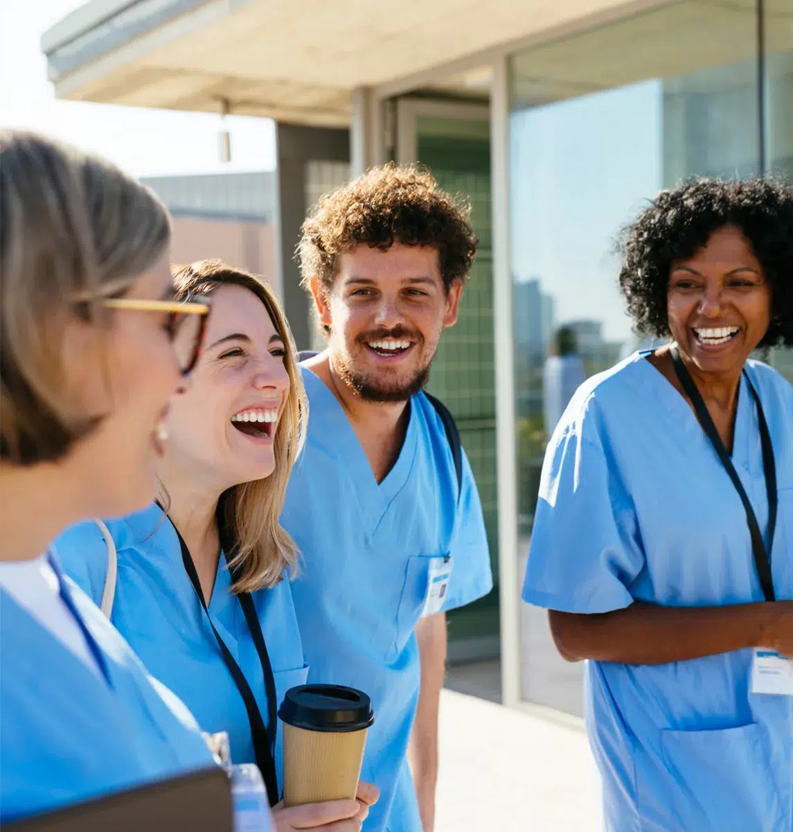 Four healthcare workers in blue scrubs smile and laugh outside a building on a sunny day, one holding a coffee cup—perfect for marketing to nurses seeking camaraderie.
