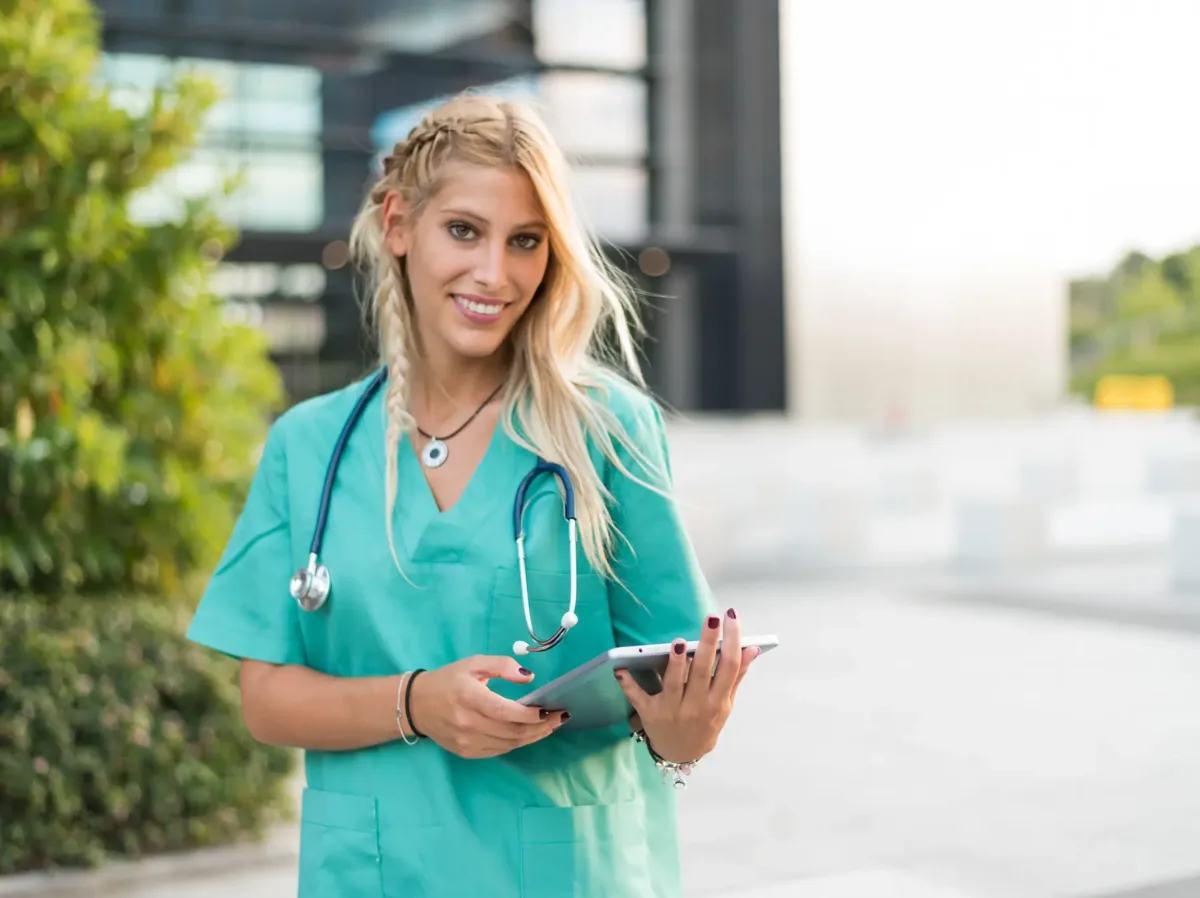 A smiling woman in green scrubs with a stethoscope around her neck holds a tablet, checking the best nurse schedule app outdoors near a modern building.