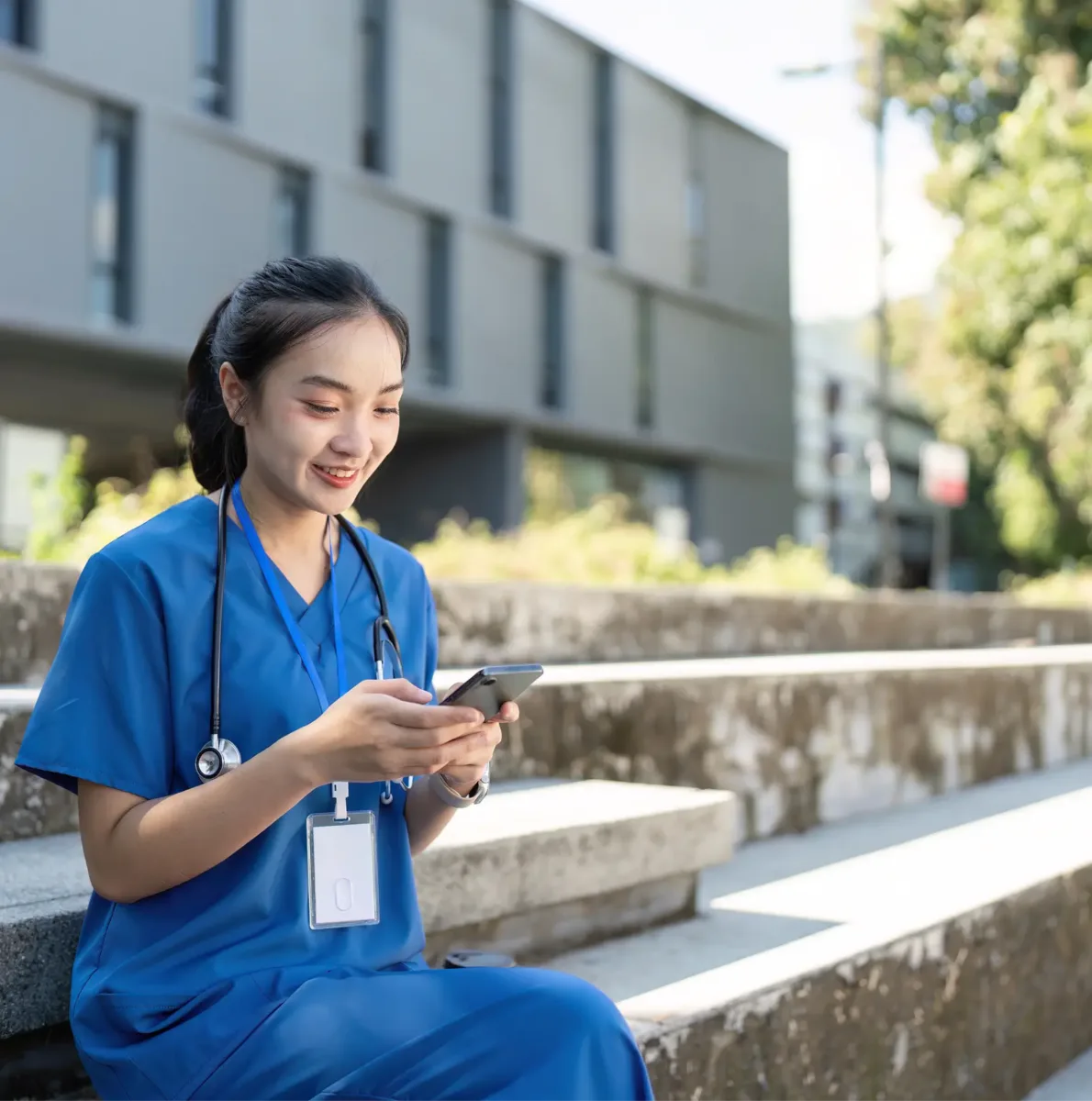 A woman in blue scrubs with a stethoscope around her neck sits on outdoor steps, smiling while using her smartphone—perhaps checking the best nursing schedule app.