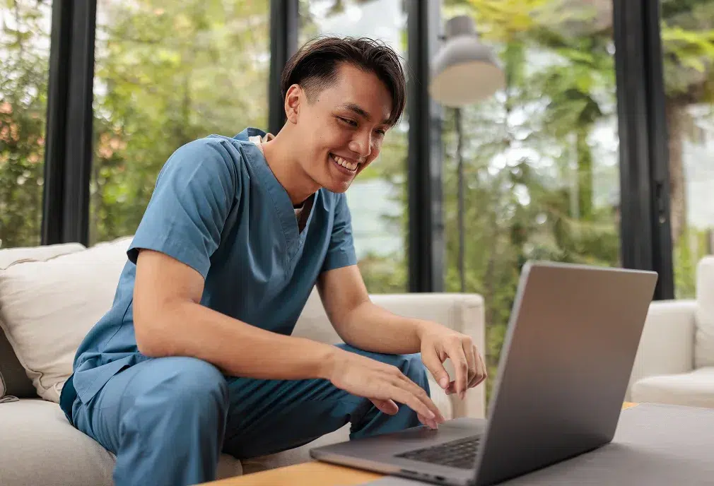 A smiling person in blue scrubs sits on a couch, typing on a laptop—perfectly capturing the essence of marketing to nurses in a bright, greenery-filled space.