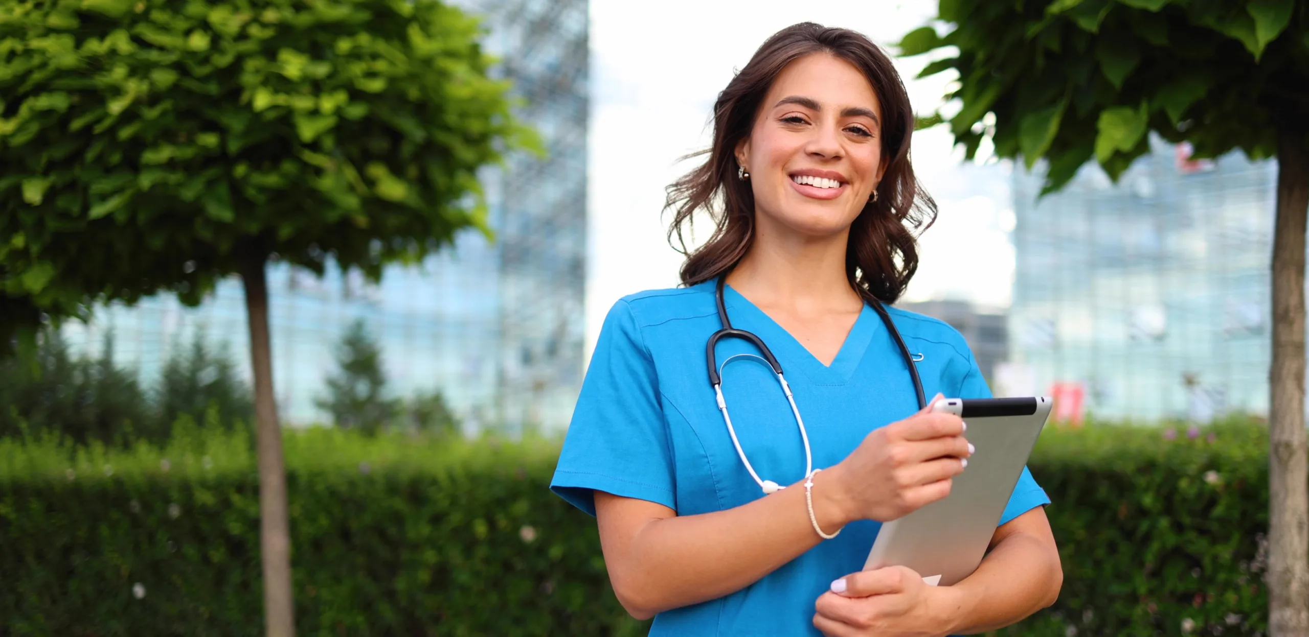 A smiling healthcare professional in blue scrubs with a stethoscope and tablet—possibly using the best nursing schedule app—stands outdoors near trees and modern buildings.