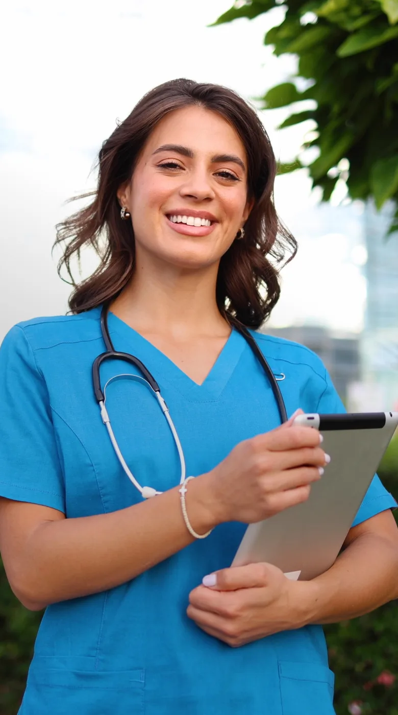 A smiling healthcare professional in blue scrubs holding a clipboard and a pen, with a stethoscope around her neck, recommends the best nursing schedule app while standing outdoors.