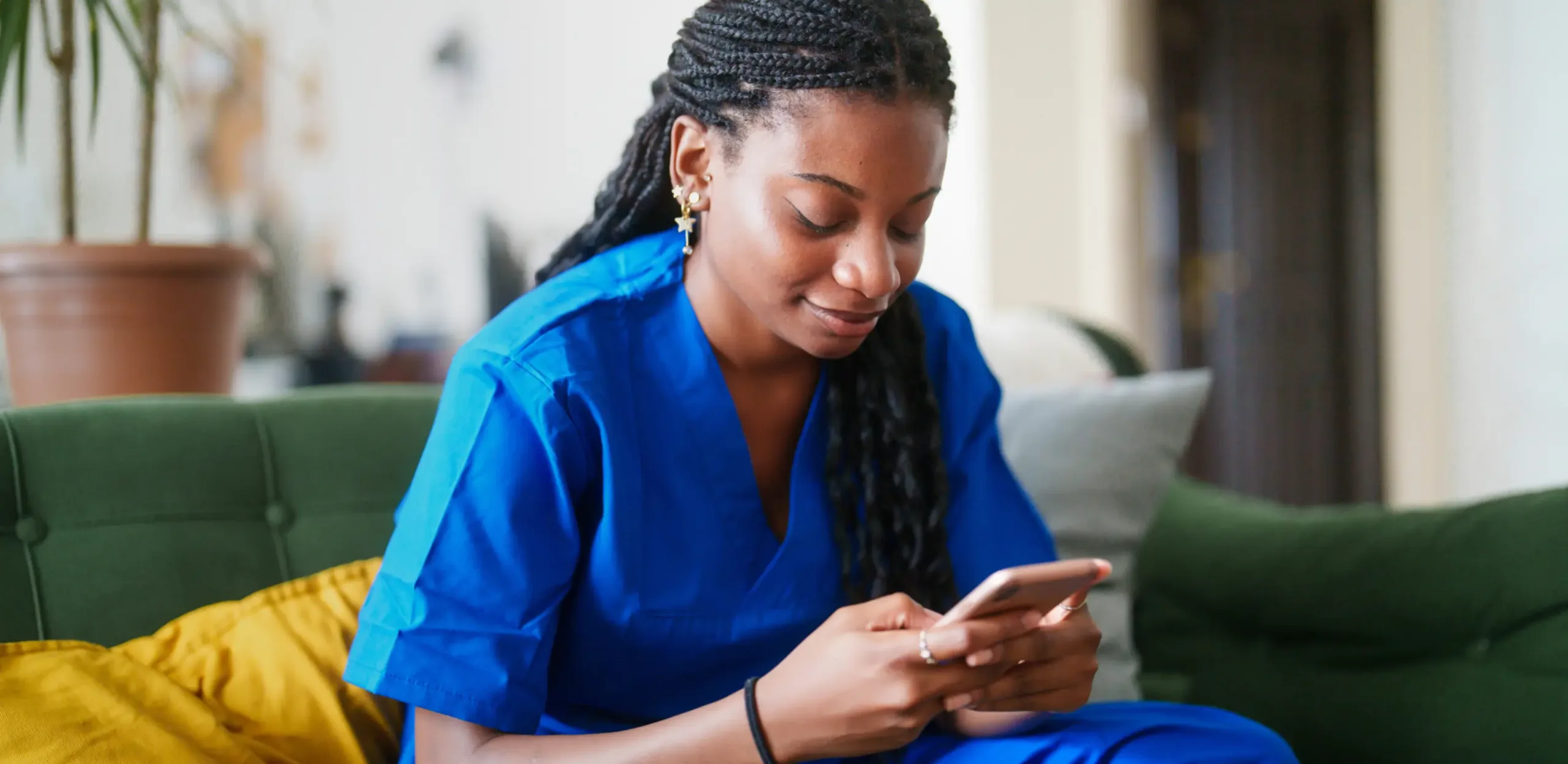 Woman in blue medical scrubs sits on a couch, smiling as she checks her nurse calendar app on her smartphone.