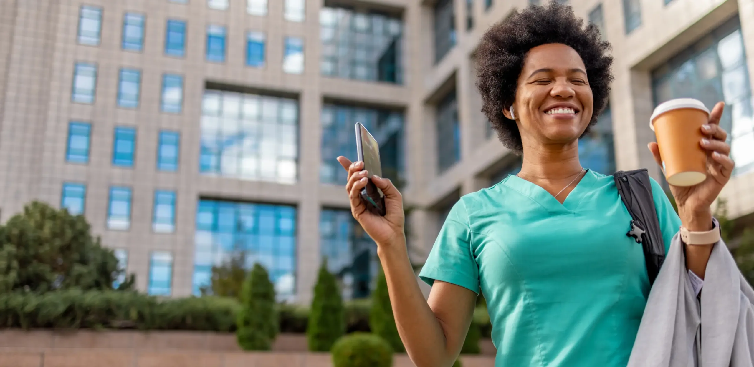 Smiling person in medical scrubs holds a phone with nurse scheduling software and a coffee cup outside a modern building, with greenery in the background.