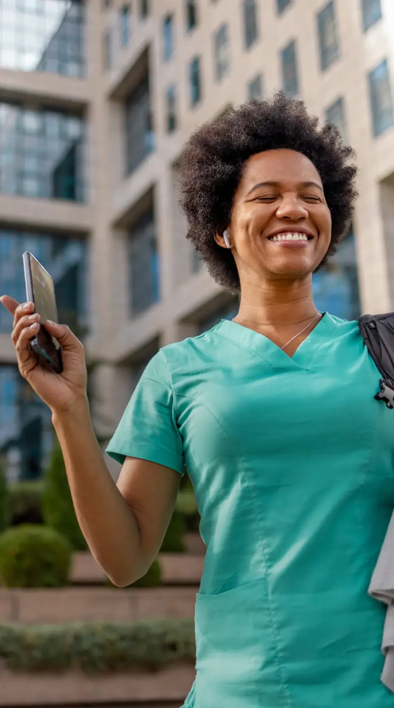 A smiling woman in green scrubs stands outdoors holding a phone, possibly checking nurse scheduling software, with a modern building in the background.