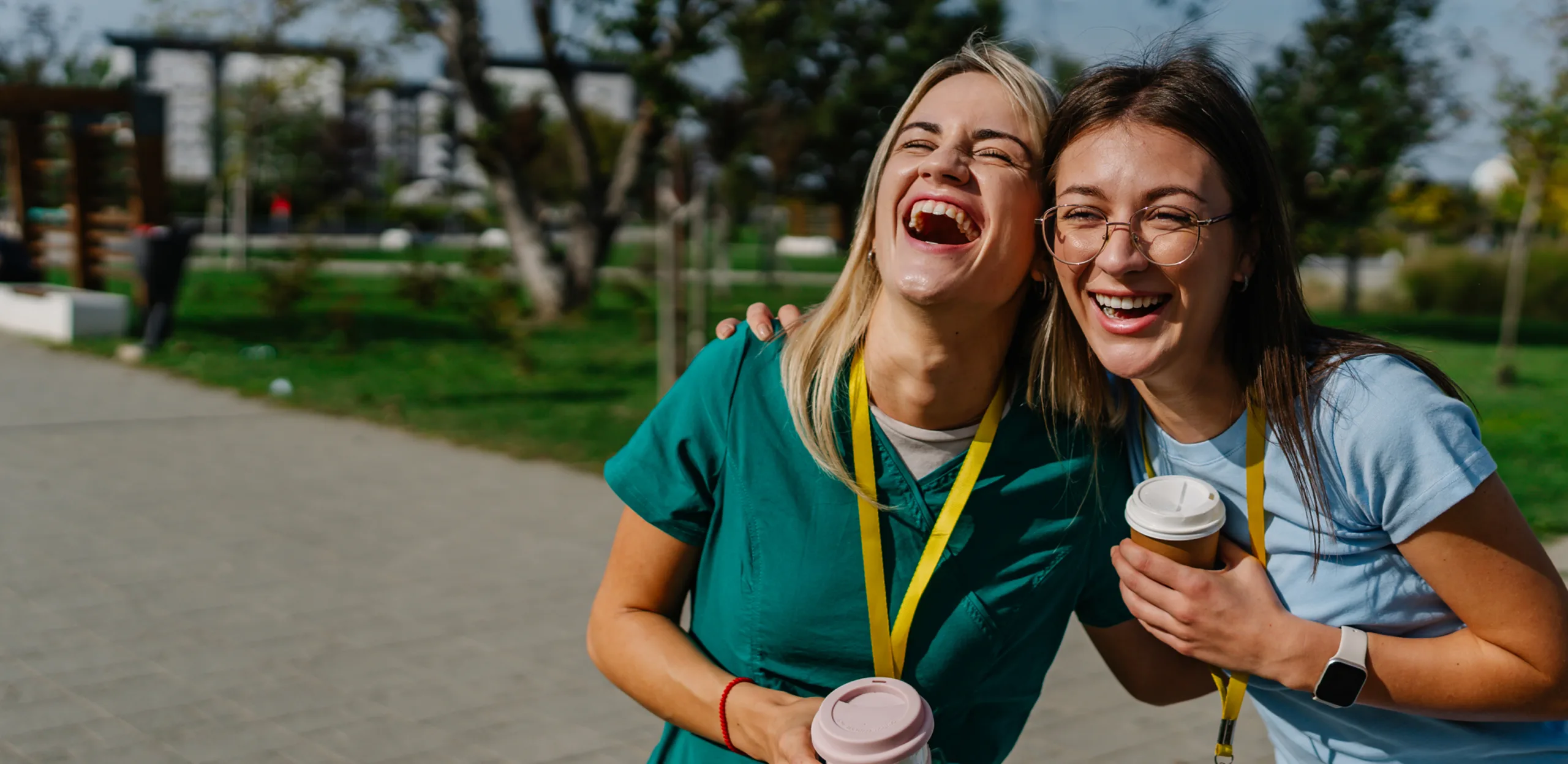 Two women with lanyards laugh and hold coffee cups outside on a sunny day, perhaps chatting about the best apps for nursing students amid the trees and buildings behind them.
