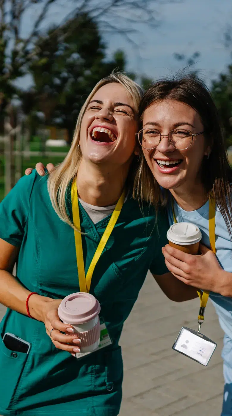 Two women in scrubs and name badges laugh together outdoors, holding coffee cups and chatting about the best apps for nursing students on a sunny day.