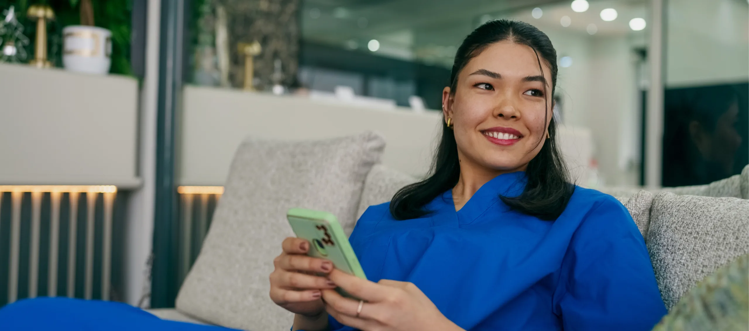 A woman in blue scrubs sits on a couch, smiling and holding her smartphone as she explores a nurse calendar app in a bright, modern indoor setting.