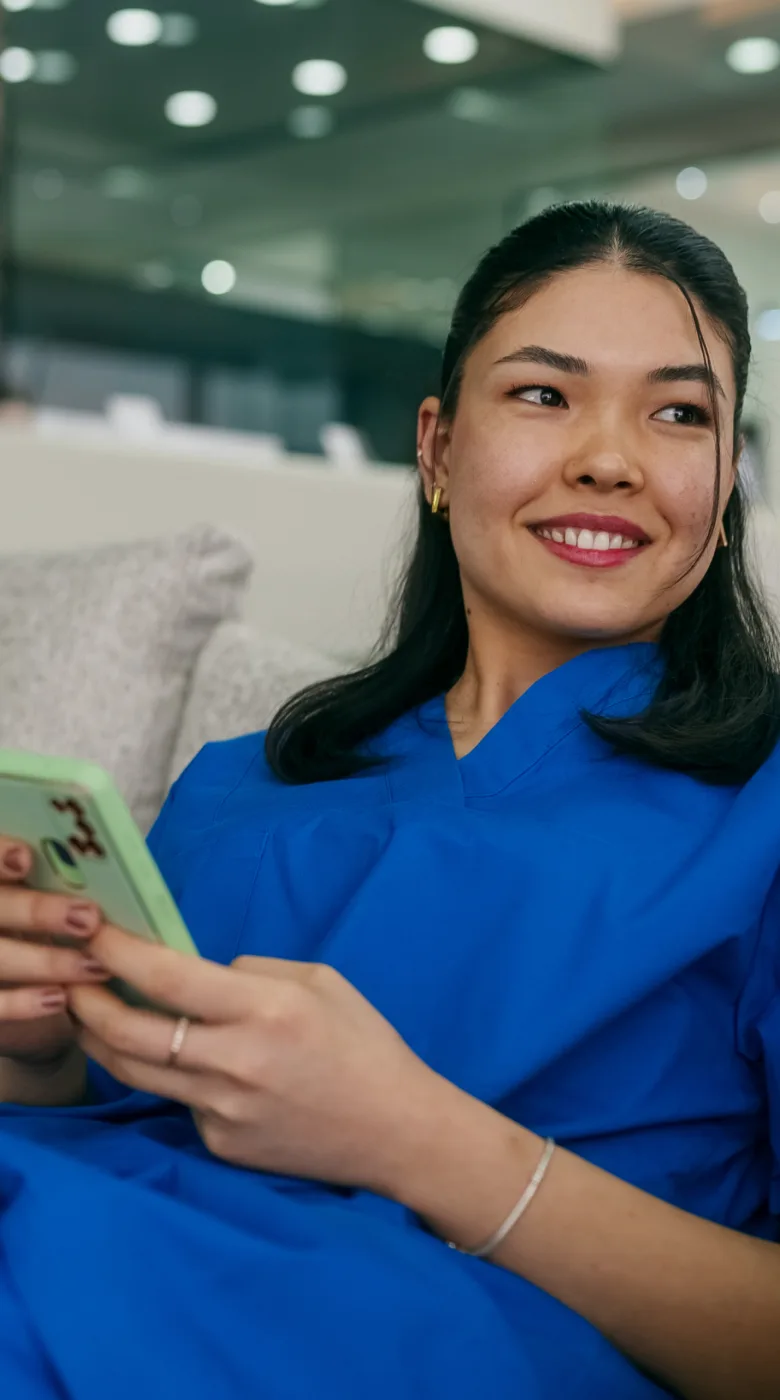 A woman in blue scrubs sits on a couch, smiling and holding her smartphone, perhaps checking her nurse calendar app in a modern, blurred indoor setting.